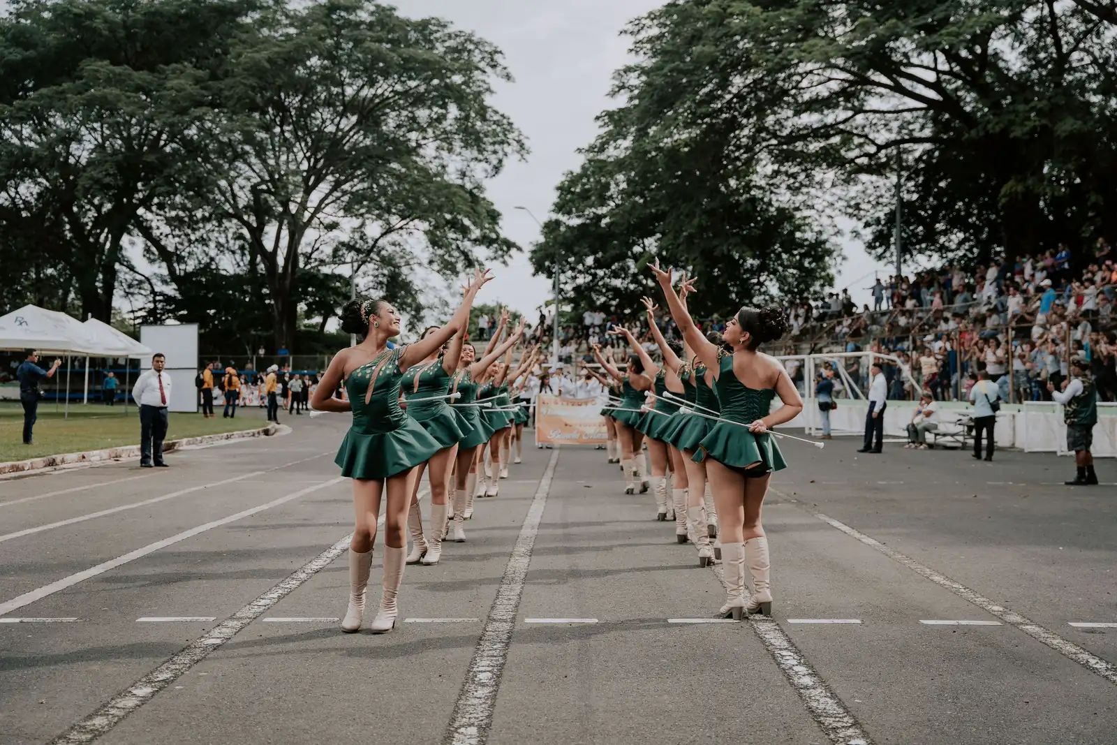 Un défilé de majorettes en tenue verte, bâton levé, sur une avenue devant le public.