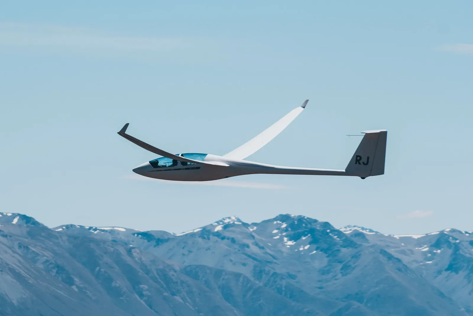 Un planeur blanc vole au-dessus d'une chaîne de montagnes enneigées sous un ciel bleu.