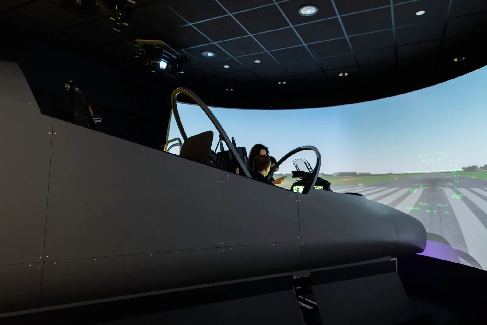 Une pilote aux commandes d'un cockpit de simulateur de vol face à un écran panoramique montrant une piste.