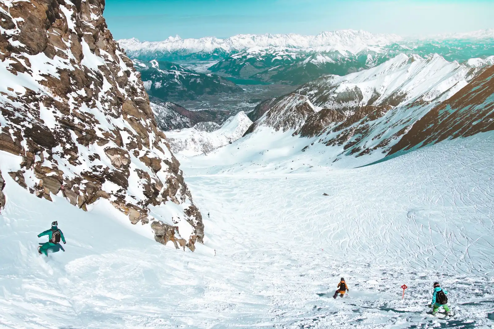 Des snowboardeurs descendent une large combe enneigée face à un panorama de sommets alpins.