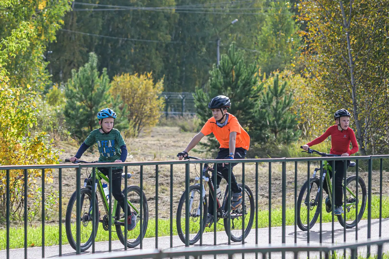 Trois enfants casqués roulent à vélo sur une piste cyclable au bord d'un parc en automne.